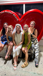 Laughing bride-to-be in a veil with four friends seated on an oversized red heart bench on a city sidewalk at night — fun bachelorette night out group photo.