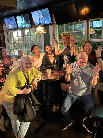 Group of adults in a lively pub around a high-top table, striking playful mock-scared poses and laughing beneath stained-glass lamps and TV screens.