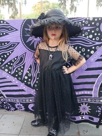 Child in Halloween vampire-witch costume wearing a wide-brim black hat, red fangs, pendant and layered black polka-dot tulle dress, posing outdoors in front of a purple celestial tapestry — spooky backyard portrait.