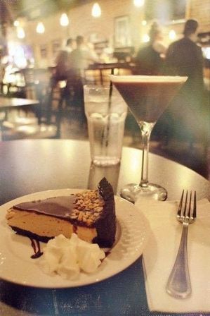 Slice of peanut butter chocolate pie with whipped cream on a plate, alongside an espresso martini and water glass on a table in a cozy bistro with warm pendant lights and blurred evening patrons