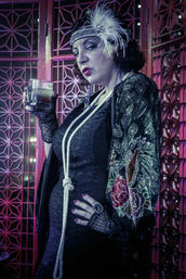 Sassy 1920s flapper in black lace dress and feathered headband holding a cocktail glass, long knotted pearl necklace and embroidered kimono sleeve, posed before a geometric art-deco screen with moody pink-purple speakeasy lighting