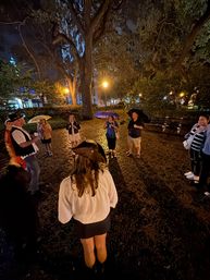 Nighttime walking tour in a city park under giant oak trees, a small group holding umbrellas in light rain gathered around a costumed guide in a tricorn hat, warm streetlamp glow and wet ground.