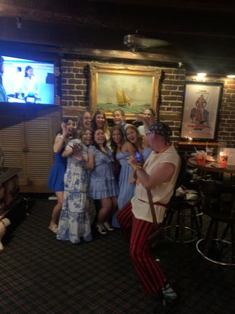 Group of women in blue and white dresses laughing and posing for a photo in a cozy brick-walled bar under a framed sailboat painting, with a costumed man in striped pants taking the picture and a TV and drinks visible