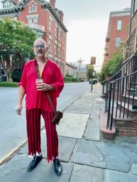 Man in a bright red shirt and red-and-black striped pants holding a drink on a stone sidewalk in a historic brick-townhouse neighborhood at dusk
