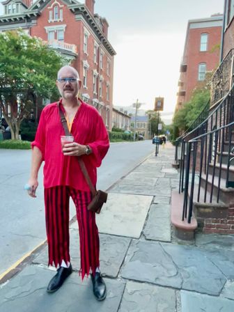 Man in a bright red shirt and red-and-black striped pants holding a drink on a stone sidewalk in a historic brick-townhouse neighborhood at dusk