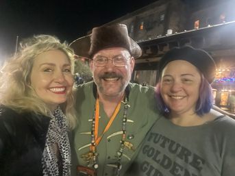 Three smiling adults posing outdoors at night in front of a lit rustic building: a man wearing a pirate-style leather hat, glasses and beaded necklaces stands between two women—one blonde with red lipstick and a patterned scarf, the other in a beret with purple-tipped hair.