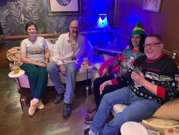 Group of four adults in festive sweaters and casual clothes seated on a plush lounge sofa in a cozy bar-style room with leafy wallpaper and blue accent lighting, holding drinks and smiling.