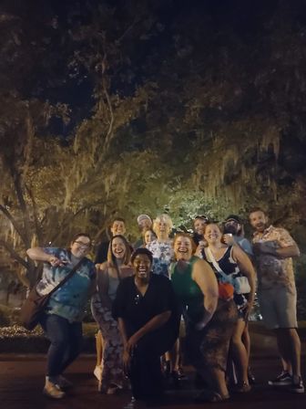 Nighttime photo of about a dozen friends smiling and laughing under moss‑draped live oak trees in a Southern park.