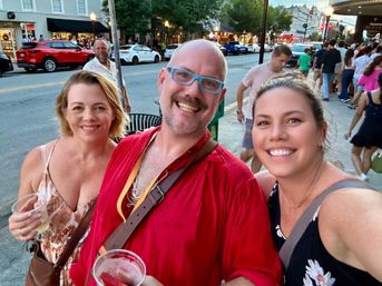 Three smiling adults taking a selfie at dusk on a busy downtown street during a summer festival, holding drinks with shops, parked cars, and a walking crowd in the background.