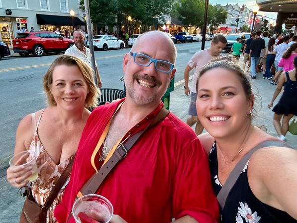 Three smiling adults taking a selfie at dusk on a busy downtown street during a summer festival, holding drinks with shops, parked cars, and a walking crowd in the background.