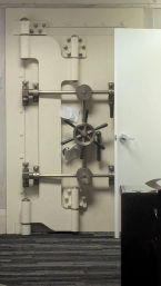 Massive cream-colored vault door with ship-style wheel and horizontal locking bars, ajar into a carpeted office space