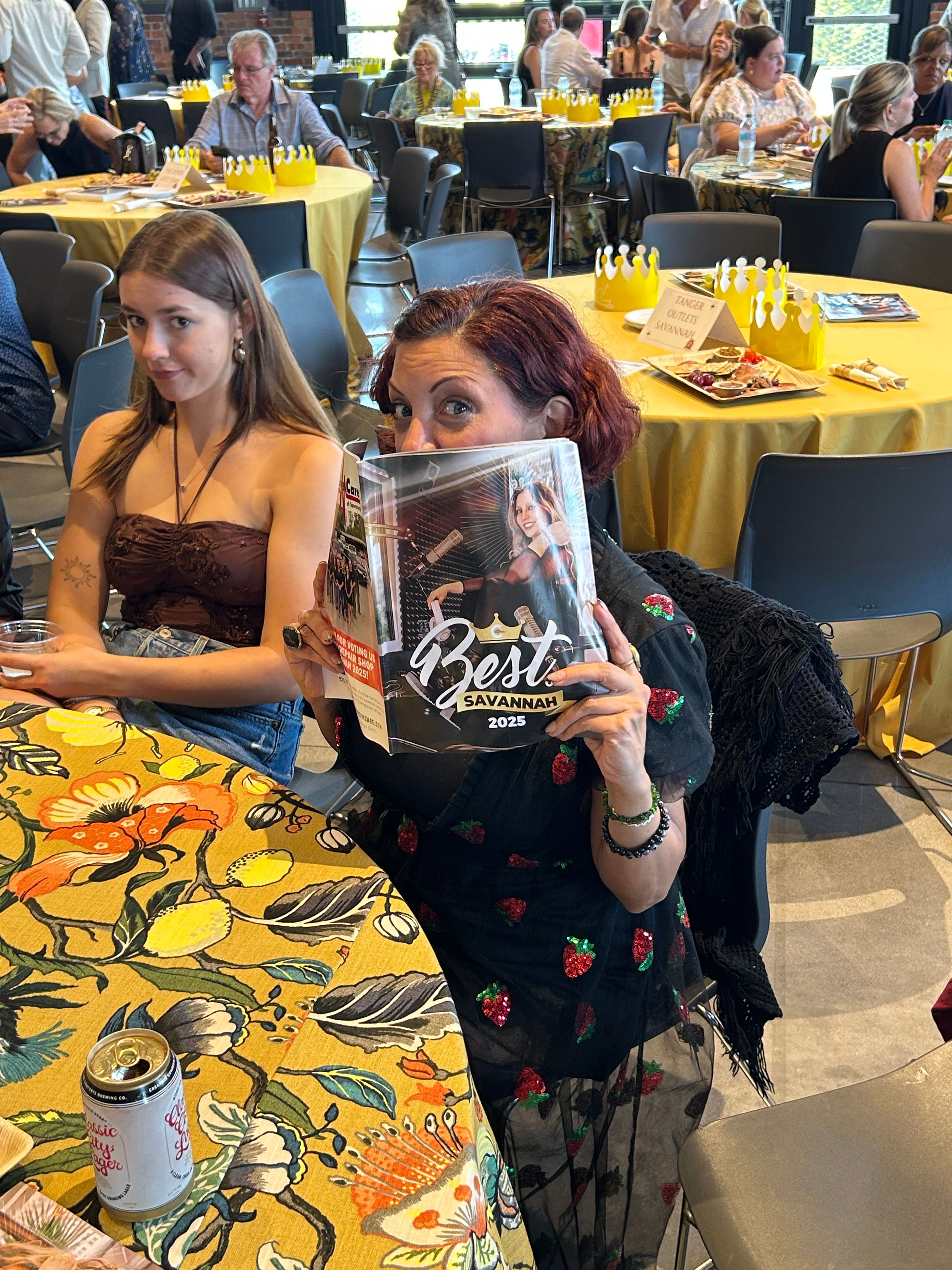 Two women at a lively Savannah awards brunch, one peeking over a 'Best Savannah 2025' magazine, round tables with yellow tablecloths and paper crown centerpieces