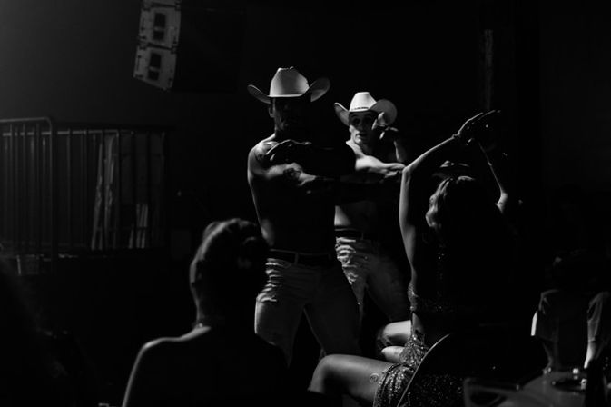 Black-and-white nightclub scene of two shirtless male dancers in cowboy hats performing for a seated audience