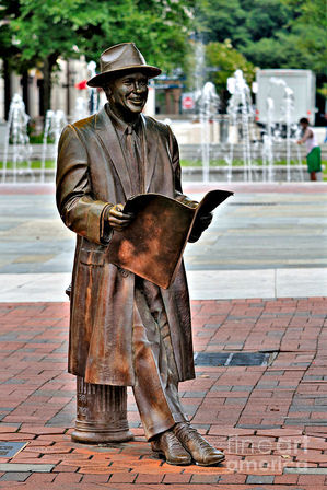 Bronze statue of a smiling man in a fedora and overcoat reading a newspaper while seated on a fire hydrant in a brick-paved downtown plaza with fountains in the background.