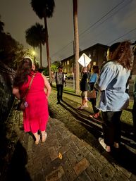 Moonlit walking tour in a historic downtown: small group on a wet brick sidewalk beneath tall palm trees, a guide gesturing by a streetlamp with old brick buildings in the background.