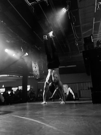 Black-and-white image of a shirtless acrobat performing a gravity-defying handstand on a dim nightclub stage under spotlights, with audience, speakers, and stage lights in the background.