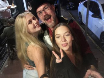 Nighttime urban selfie of three friends on a sidewalk: two smiling women and a bearded man in a hat laughing, brunette making a peace sign with parked cars and passersby in the background.