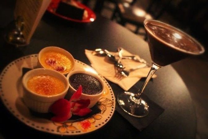 Three ramekins of dessert—two caramelized custards and one chocolate custard—on a decorative plate with orchid petals, paired with a chocolate cocktail in a martini glass on a dimly lit table.