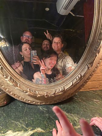 Group selfie of five people in an ornate round mirror at a dimly lit bar — two flashing peace signs, tattooed arm visible, green marble countertop in the foreground.