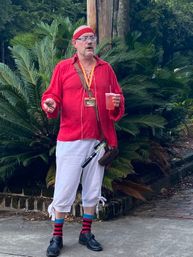 Person in a red pirate-style costume — red shirt and headband, blue glasses, white cropped pants and red-and-black striped socks — holding a pink drink and phone with a satchel and prop sword, standing on a sidewalk beside lush palm-like plants