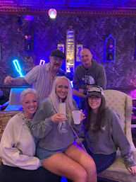 Group of five friends smiling and holding drinks in a neon-lit indoor lounge with purple-blue lighting, vintage patterned wallpaper and cozy seating — casual night out.