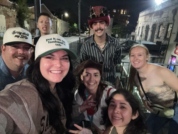 Group selfie of seven friends on an urban balcony at night, smiling and wearing playful hats including a steampunk top hat and cowboy hat, holding drinks and enjoying nightlife