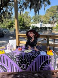 Smiling tarot reader in a red-and-black wide-brimmed hat seated behind a purple celestial tarot cloth at an outdoor market, displaying tarot cards, cash, a lantern and a small pumpkin beneath oak trees draped with Spanish moss.