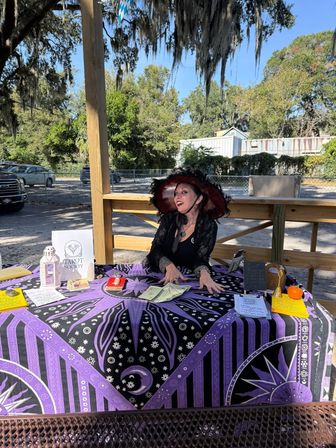 Smiling tarot reader in a red-and-black wide-brimmed hat seated behind a purple celestial tarot cloth at an outdoor market, displaying tarot cards, cash, a lantern and a small pumpkin beneath oak trees draped with Spanish moss.