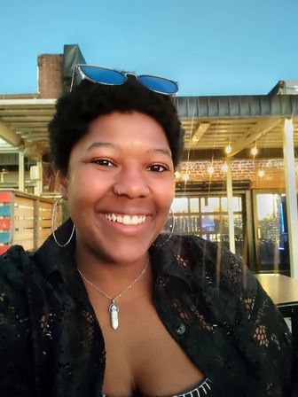 Smiling person with short curly hair, hoop earrings, nose ring and pendant necklace, sunglasses on their head, seated at a covered outdoor patio with hanging string lights at dusk.
