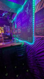 Vibrant neon-lit bar interior with teal LED-trimmed chalkboard craft beer menu, purple zebra-pattern wall, wooden counter and bar stools
