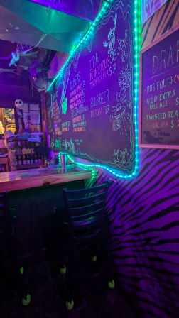 Vibrant neon-lit bar interior with teal LED-trimmed chalkboard craft beer menu, purple zebra-pattern wall, wooden counter and bar stools