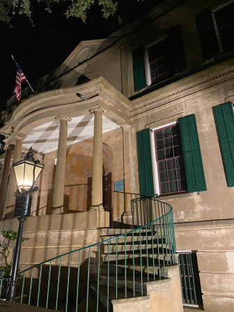 Night view of a historic colonial-style house with a columned porch, curved exterior spiral staircase, green shutters, a lit street lamp and an American flag.