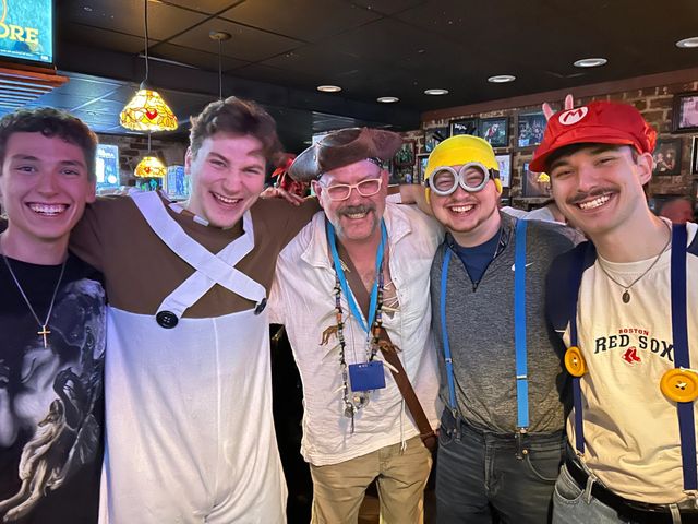 Five people in playful costumes — pirate, Mario hat, minion goggles, and a white onesie — smiling together at a lively pub costume party with stained-glass pendant lamps and framed photos on a brick wall.