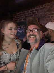 Lively nightlife scene in a brick‑walled bar: a laughing man in a tricorn hat and glasses poses beside a smiling woman in a newspaper‑print dress with visible tattoos.