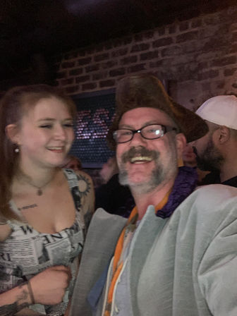Smiling man wearing glasses and a tricorn hat beside a laughing young woman with a tattoo, posing for a selfie inside a dim, brick-walled bar during a lively night out.