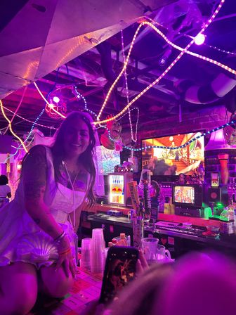 Smiling woman in a white outfit perched on a neon-lit basement bar counter surrounded by multicolored LED rope lights, drink taps, plastic cups and retro TVs.