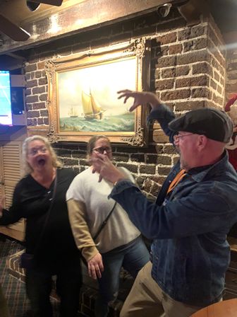 Three adults joking in a cozy brick-walled pub under a framed sailboat painting, a man striking a playful mock-scary pose while two women react and laugh.