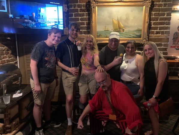 Seven friends posing in a cozy brick-walled pub under a framed sailboat painting, some holding drinks and one person in a red costume crouching in front — casual night out.