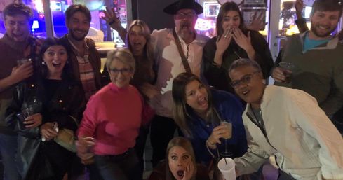 Group of people posing with surprised expressions and drinks in hand at a lively downtown bar night with neon lights.