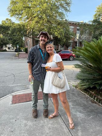 Smiling young couple posing on a sunny historic downtown corner with brick rowhouses and palm fronds — woman in a white sundress holding a drink and beige shoulder bag, man in a striped shirt, green pants and a novelty hat holding a beverage.