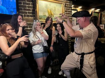 Costumed pirate performer playfully miming a scare while a group of women laugh and react with drinks in hand inside a cozy brick-walled pub beneath a framed sailboat painting.