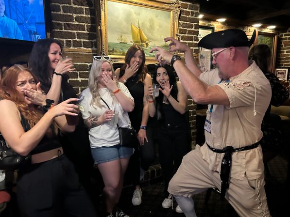 Costumed pirate performer playfully miming a scare while a group of women laugh and react with drinks in hand inside a cozy brick-walled pub beneath a framed sailboat painting.