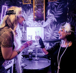 Two women in 1920s flapper outfits toast with champagne flutes in a purple-lit speakeasy-style lounge, wearing feathered headbands, long pearls, and vintage wallpaper backdrop.
