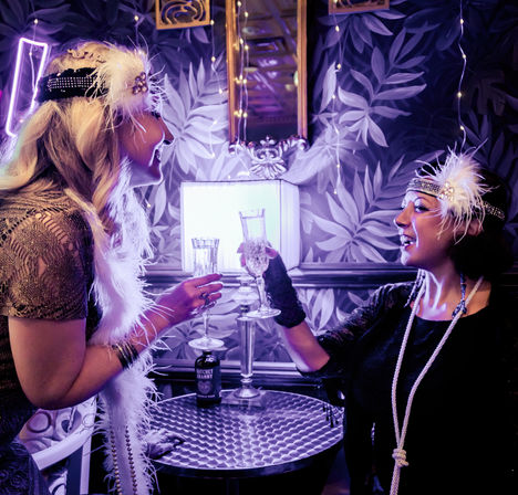 Two women in 1920s flapper outfits toast with champagne flutes in a purple-lit speakeasy-style lounge, wearing feathered headbands, long pearls, and vintage wallpaper backdrop.