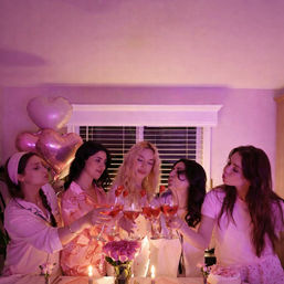 Five friends in pink pajamas toasting glasses of rosé with strawberry garnishes at a pink-themed indoor girls’ night, surrounded by heart-shaped balloons, candles, flowers and a small cake.