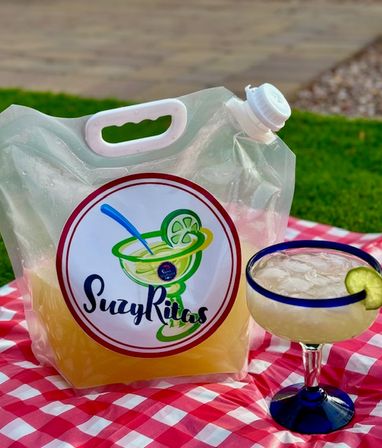 Margarita mix in a clear to-go pouch with spout and cocktail logo next to a blue-rimmed margarita glass with lime wedge, on a red-and-white checkered picnic blanket in a grassy backyard.