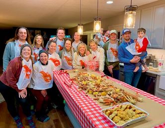 Smiling family and friends in a home kitchen wearing lobster bibs, gathered around a red-checked table piled with crab legs, corn and potatoes from a seafood boil under pendant lights
