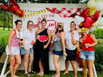 Six friends posing at a summer backyard party photo booth on a lawn, holding drinks and playful props in front of a “Let the Good Times” backdrop with red crab balloons and yellow string lights.
