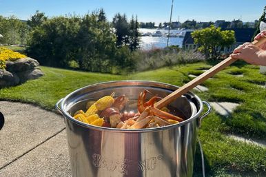 Stainless steel pot of steaming seafood boil with lobster, crab legs, corn and potatoes being stirred with a wooden paddle in a sunny waterfront backyard overlooking a marina.