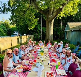 Outdoor summer backyard lobster boil under a large tree — long red-checkered table with family and friends eating lobsters, paper plates and red cups in a suburban yard.